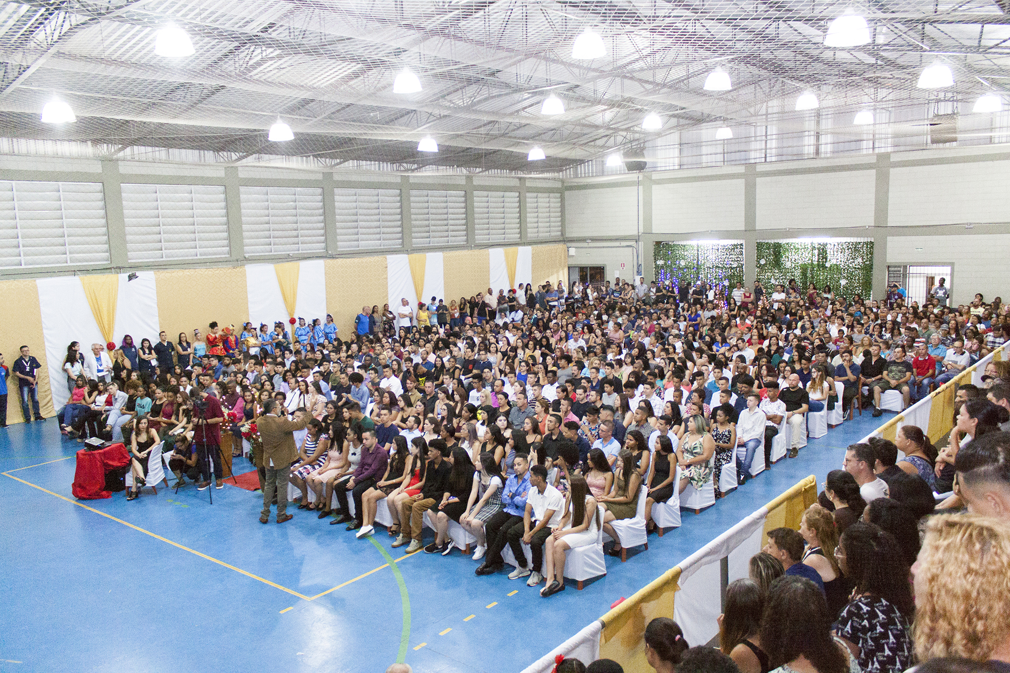 Convidados sentados em cadeiras brancas lotando o ginásio. Na frente o mestre de cerimônias faz uma apresentação.