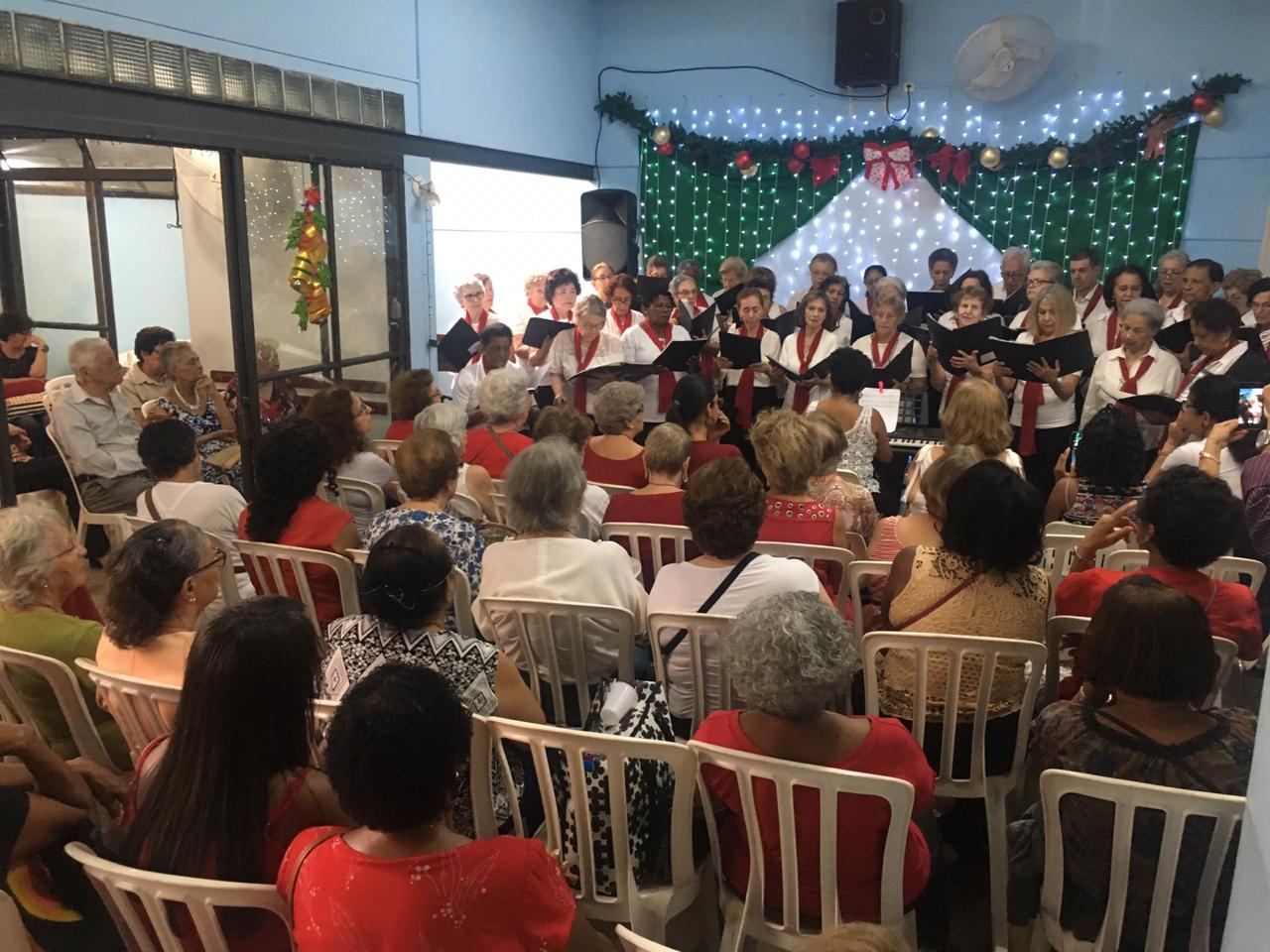 Vários idosos sentados de costas assistindo a apresentação do coral.