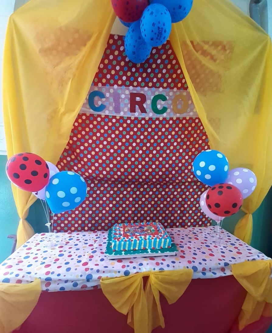 Bolo dos aniversariantes do mês em um cenário colorido enfeitado com balões dos dois lados com um letreiro escrito “Circo” em cima.