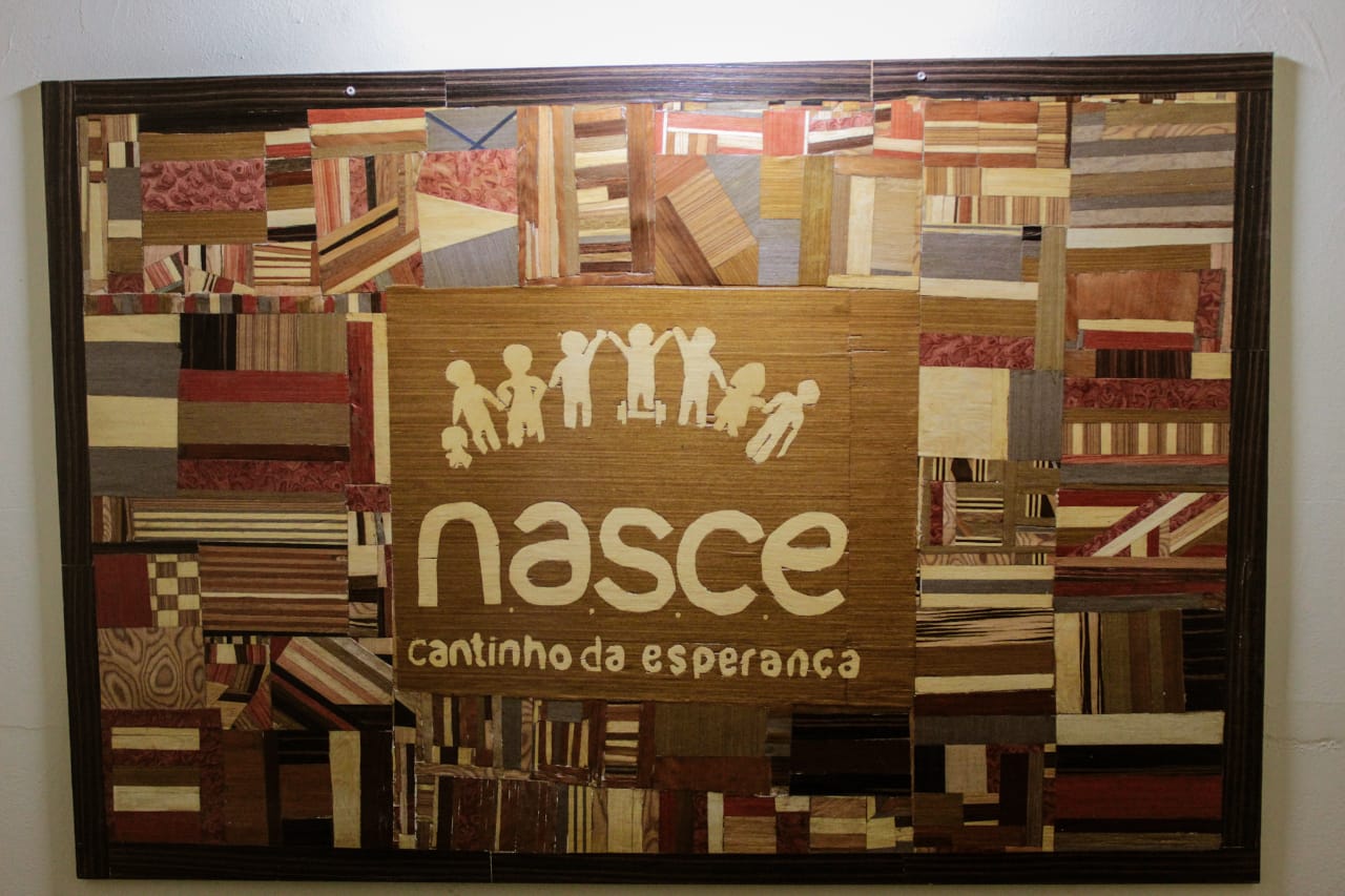 Um quadro feito pelos alunos utilizando a técnica de marchetaria, no centro o logo NASCE Cantinho da Esperança.  