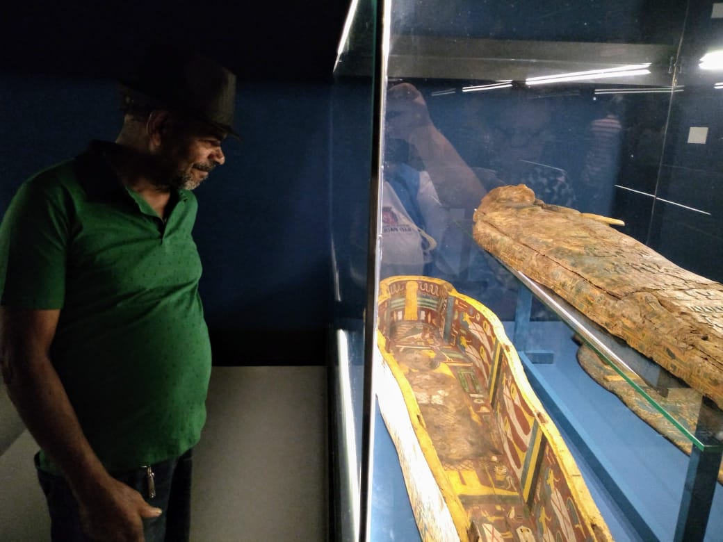 Um homem de blusa verde observa um sarcófago antigo.