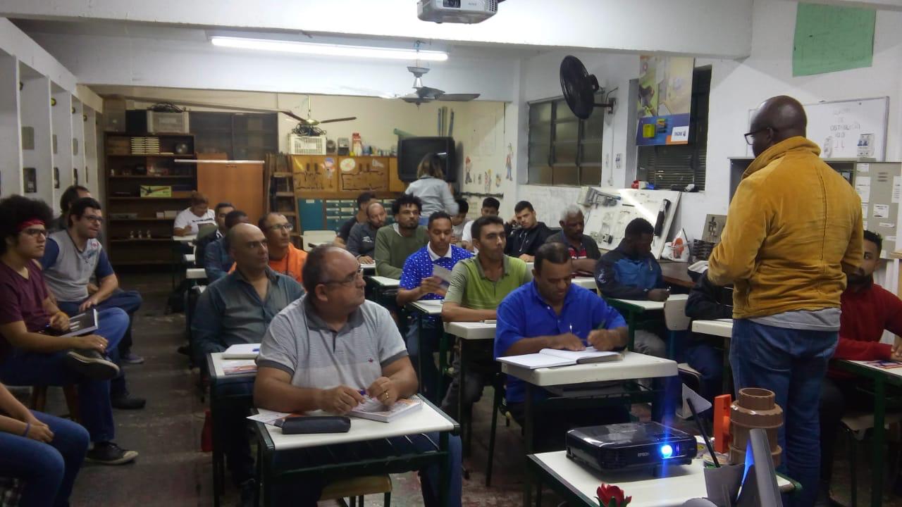 Sala de aula onde aparecem alunos sentados em carteiras escolares enfileiradas. Eles observam ao homem que está a frente da sala palestrando com auxílio de material visual projetado na parede. 