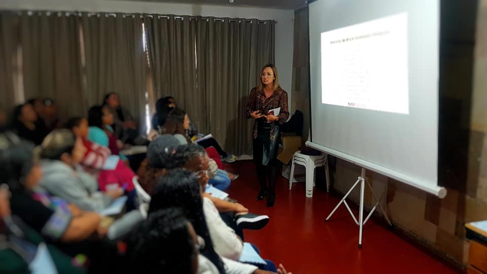 Cerca de 20 mulheres aparecem sentadas em cadeiras em uma sala, assistindo a apresentação de uma mulher em pé, do lado de uma tela que reflete imagens de um projetor. A foto foi capturada do ponto de vista lateral da sala.