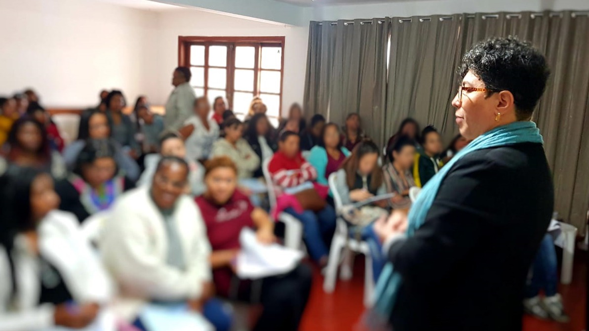 Cerca de 60 mulheres em uma sala sentadas em cadeiras assistindo a apresentação de mulher em pé, do lado de uma tela que reflete imagens de um projetor. A foto foi capturada do ponto de vista frontal da sala, ou seja, onde está a mulher em pé.