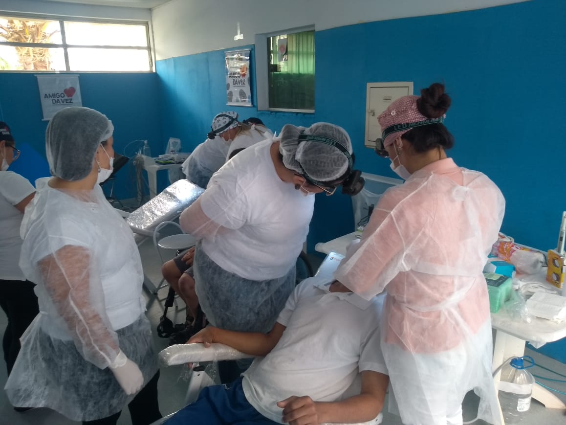 em um dos espaços da instituição Lar Ternura, mais de dez pessoas na foto. Em uma maca, uma pessoa com deficiência é atendida pela equipe de dentistas, mais de 3 pessoas ao redor.