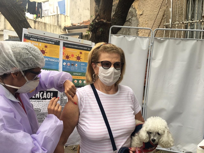 #PraCegoVer: Uma enfermeira com touca, jaleco e máscara, na cor branca aplica uma injeção no braço de uma outra senhora. A outra senhora está usando óculos de sol, máscara no rosto e segura um cachorrinho poodle. 