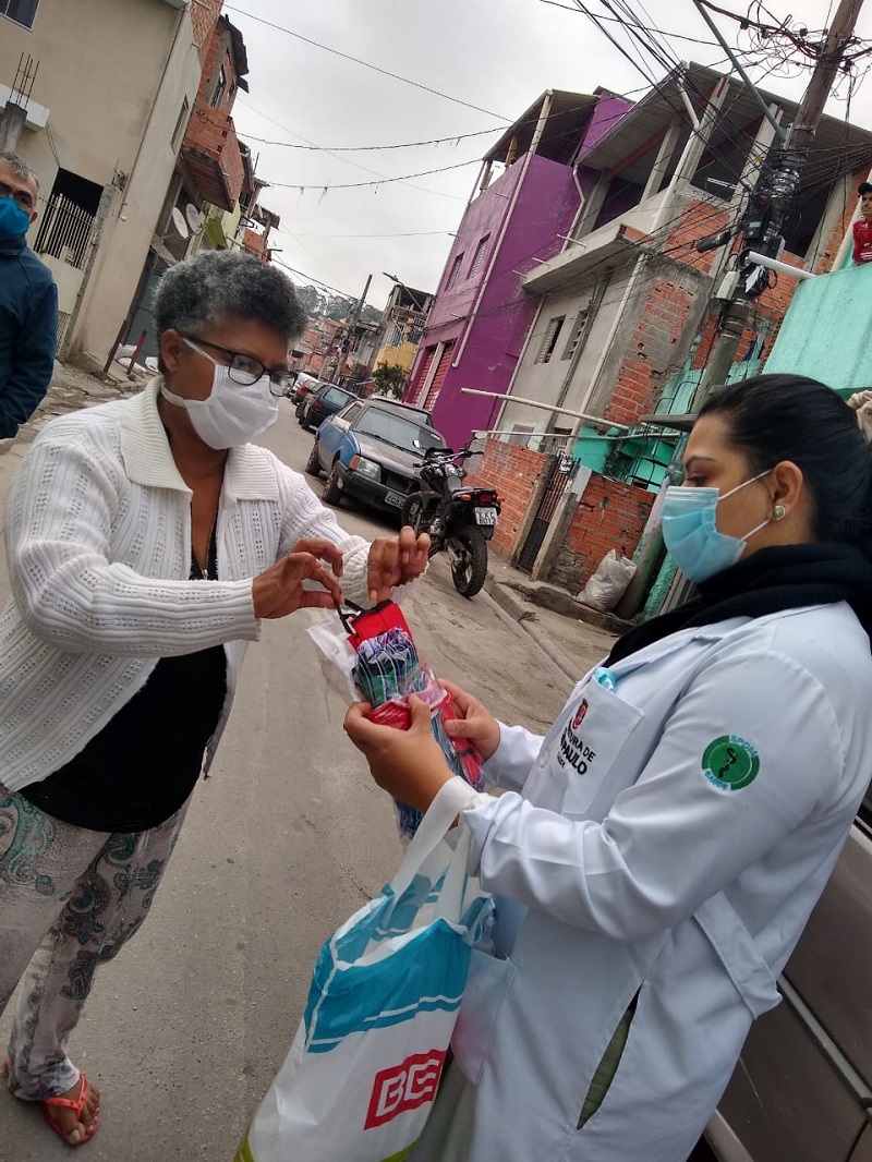 #PraCegoVer: Uma enfermeira vestindo um jaleco branco e máscara de proteção no rosto, entregando uma máscara para uma senhora na rua. A senhora usa óculos de grau, usa máscara de proteção no rosto.