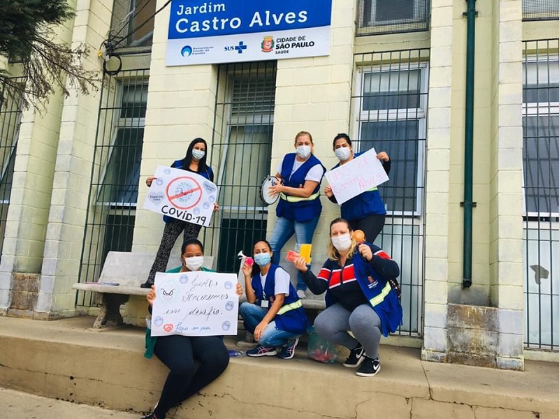 #PraCegoVer: Seis agentes de saúde segurando cartazes com mensagens contra o coronavírus. Elas estão usando coletes azuis e máscaras de proteção no rosto. Elas estão em frente à placa da UBS Jardim Castro Alves.