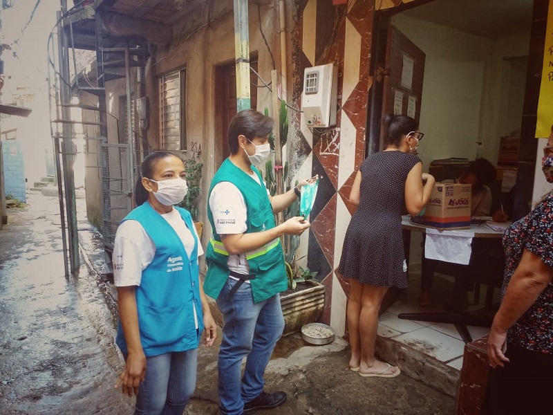 #PraCegoVer: Dois agentes de saúde. Um veste colete verde, e a outra veste um colete azul. Os dois usam máscara de proteção no rosto. Eles estão auxiliando na entrega de cestas básicas.