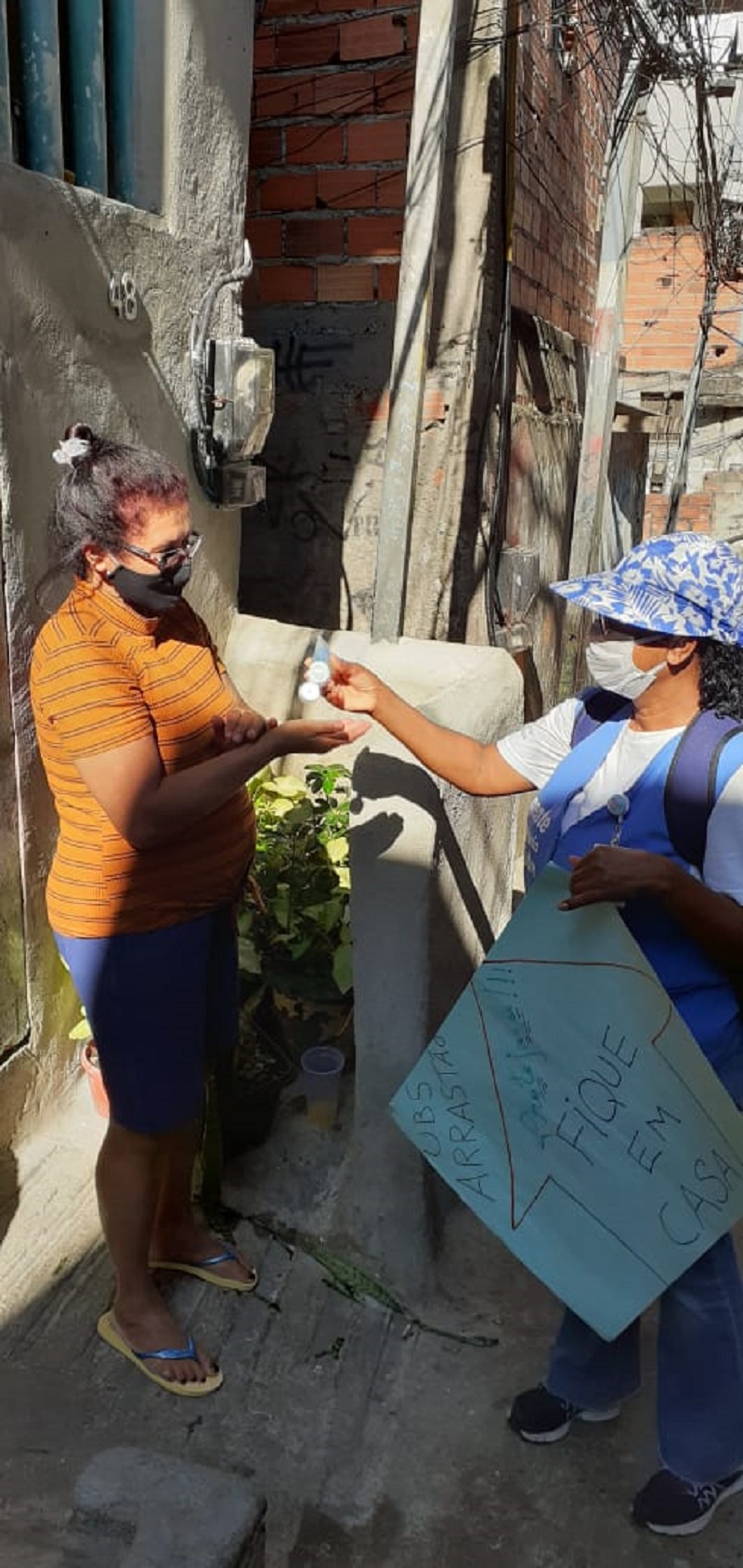 #PraCegoVer: Agente de saúde coloca álcool em gel nas mãos de uma mulher. A agente usa um colete azul, a outra mulher veste uma camiseta laranja. As duas usam máscaras de proteção no rosto.