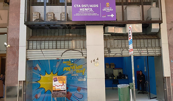 #pracegover foto da fachada do CTA DST/AIS Henfil, mostrando a placa nova, com a cor violeta. 