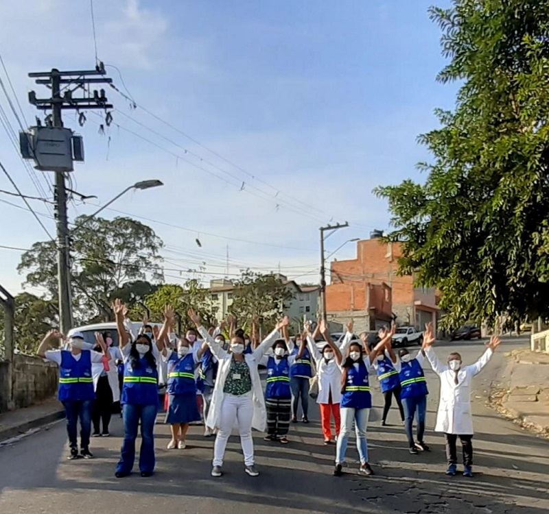 #PraCegoVer: Funcionárias da UBS estão reunidas no meio de uma rua. São 15 mulheres de braços levantados, 4 mulheres usam jalecos brancos e as outras usam jaleco na cor azul.