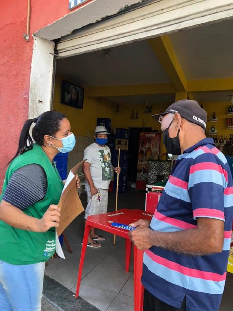 #PraCegoVer: Mulher conversando com dois homens. Ela veste um colete verde, e mostra um cartaz para um dos homens sobre prevenção do coronavírus. Eles estão na porta de um bar.