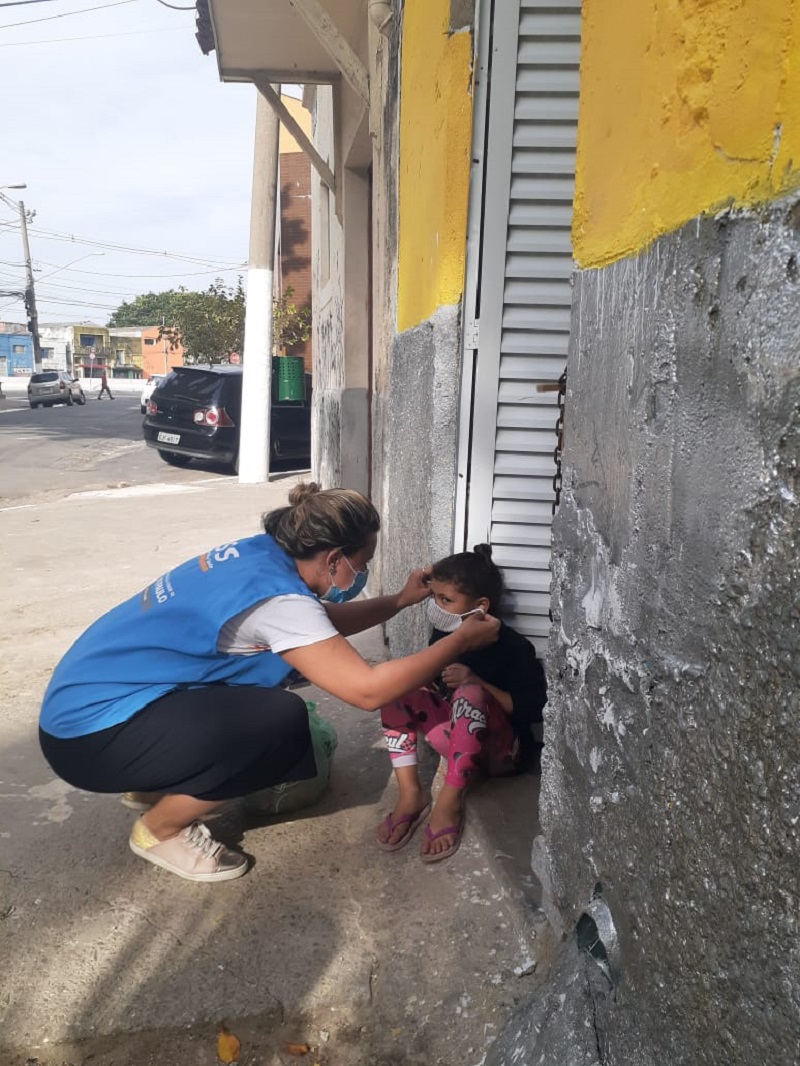 #PraCegoVer: Uma criança está sentada no chão, uma mulher se abaixa na altura da criança para colocar a máscara em seu rosto. A mulher está usando um colete azul, a criança usa uma calça rosa e blusa preta.