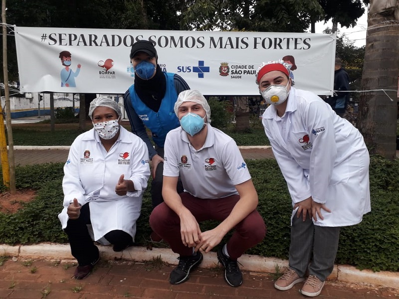 #PraCegoVer: Quatro pessoas abaixadas para tirar foto. Duas mulheres usam jaleco branco, uma usa colete azul e um homem usa camiseta branca. Todos estão com máscara de proteção no rosto. Eles estão em frente a um banner de prevenção contra o coronavírus.