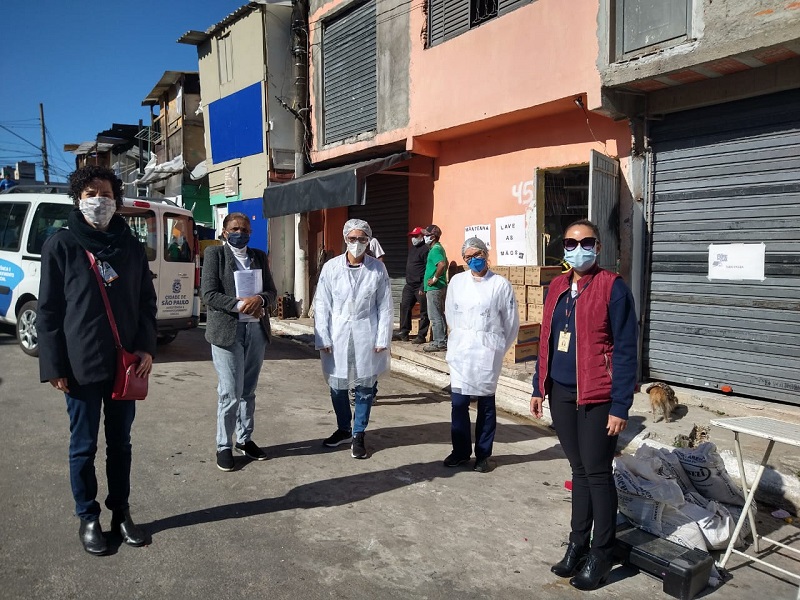 #PraCegoVer: Cinco mulheres em pé, usando máscaras de proteção no rosto. Elas estão pousando para foto. De fundo temos algumas casas.