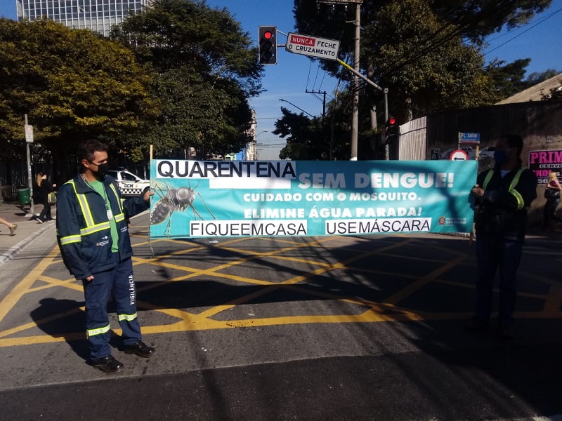 #PraCegoVer: Dois homens segurando um banner com a frase "Quarentena sem dengue. Cuidado com o mosquito. Elimine água parada. Fique em casa. Use máscara". Eles estão em uma faixa de pedestres. 