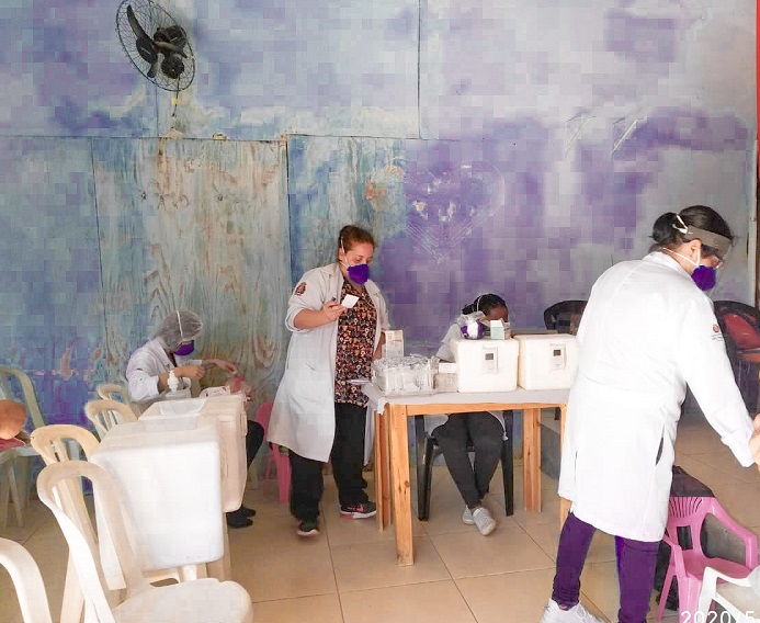 #PraCegoVer: Há quatro mulheres na foto, elas usam toucas e jalecos na cor branca, máscaras na cor roxa, elas estão organizando vacinas dentro de uma caixa de isopor. É uma sala com muitas cadeiras, todas na cor branca