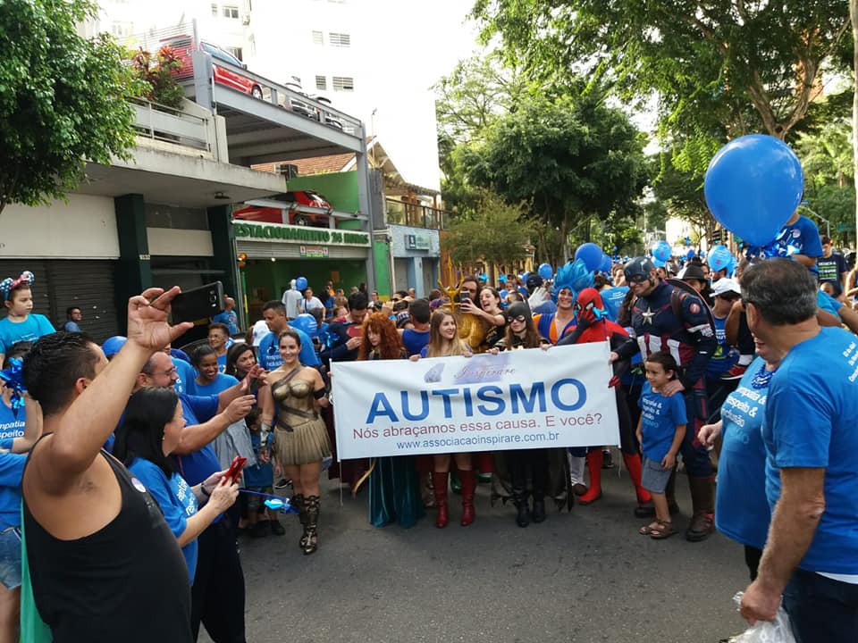 aproximadamente 60 pessoas na foto, na Avenida Paulista. As pessoas usam camisetas na cor azul e balões também na cor azul. Quatro pessoas na frente, seguram uma faixa escrita: Autismo.