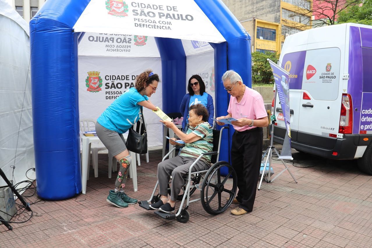  em frente da tenda da SMPED, quatro pessoas. Equipe da SMPED realiza atendimento de uma senhora em uma cadeira de rodas. 