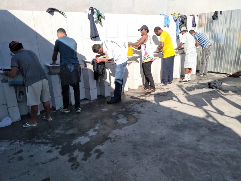 #PraCegoVer: Sete homens enfileirados em frente a tanques com torneiras. Eles estão lavando suas roupas.