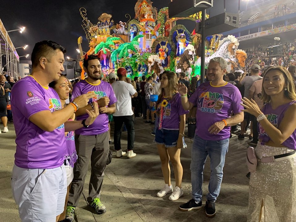 Mais de cinco pessoas na foto, surdos no camarote acompanham o desfile. Escola de samba passa na avenida ao fundo, destaque para um dos carros. 