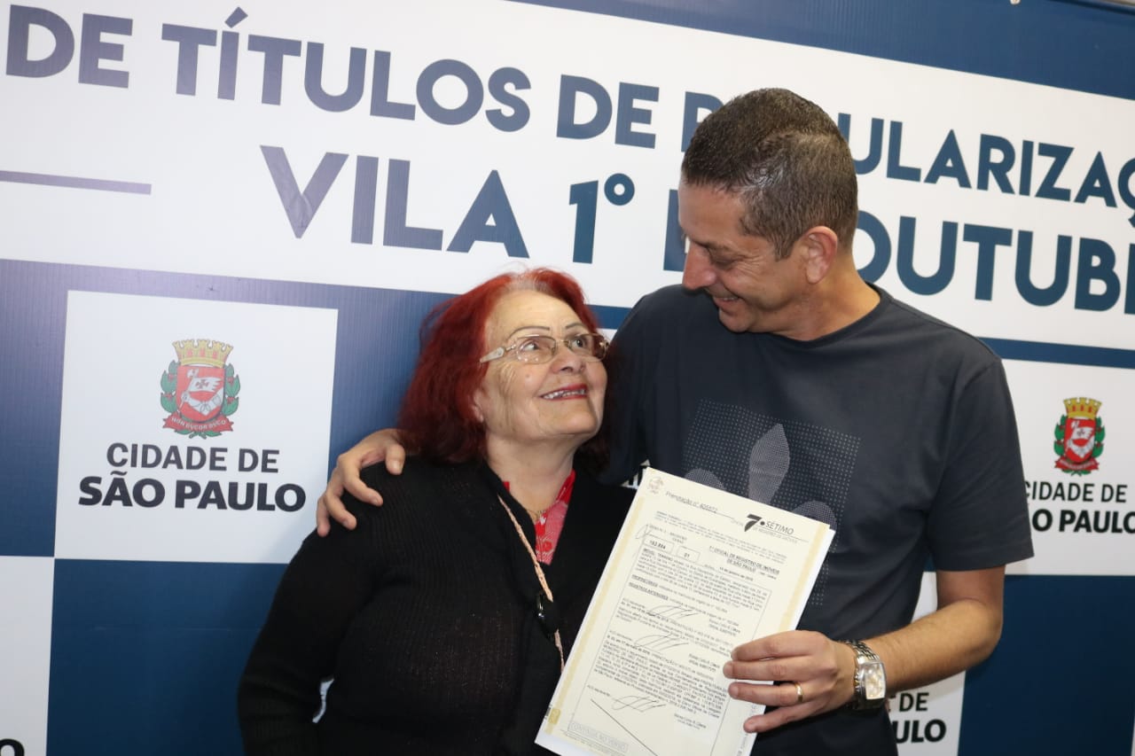 Na imagem, a foto do Secretário Municipal de Habitação, João Farias, ao lado de uma munícipe que recebe seu termo de quitação em mãos.