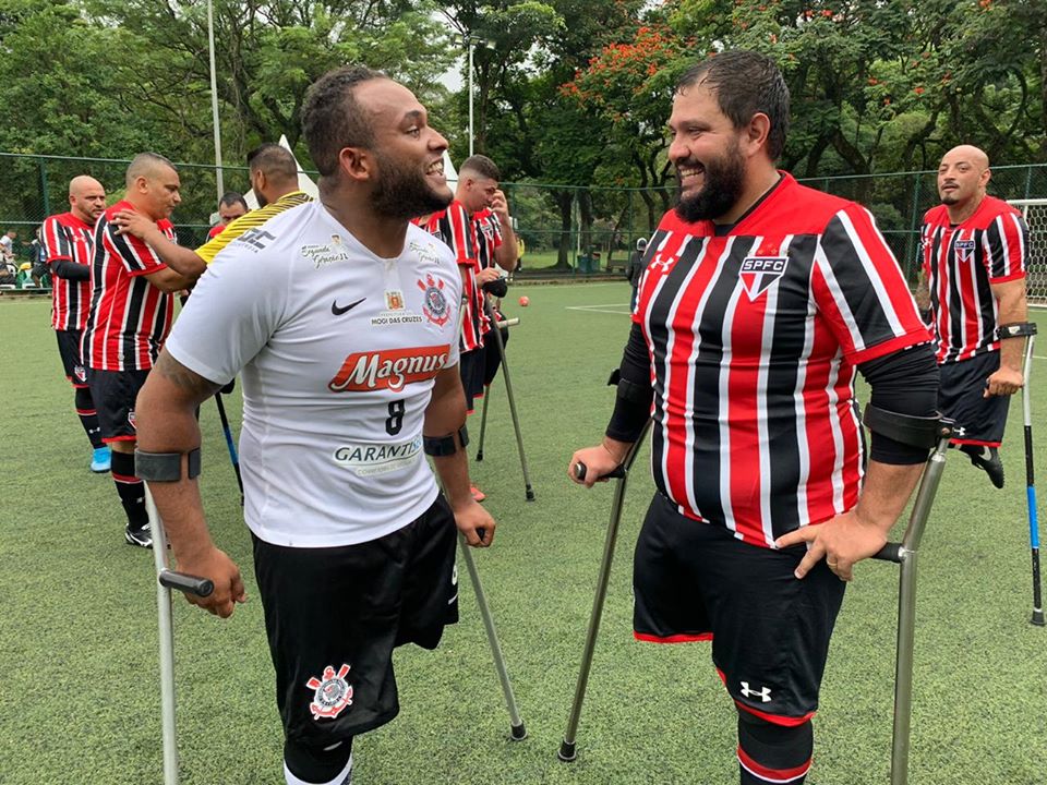 mais de cinco pessoas na foto, dois jogadores um do Corinthians e outro do São Paulo rindo um para o outro no campo de futebol.
