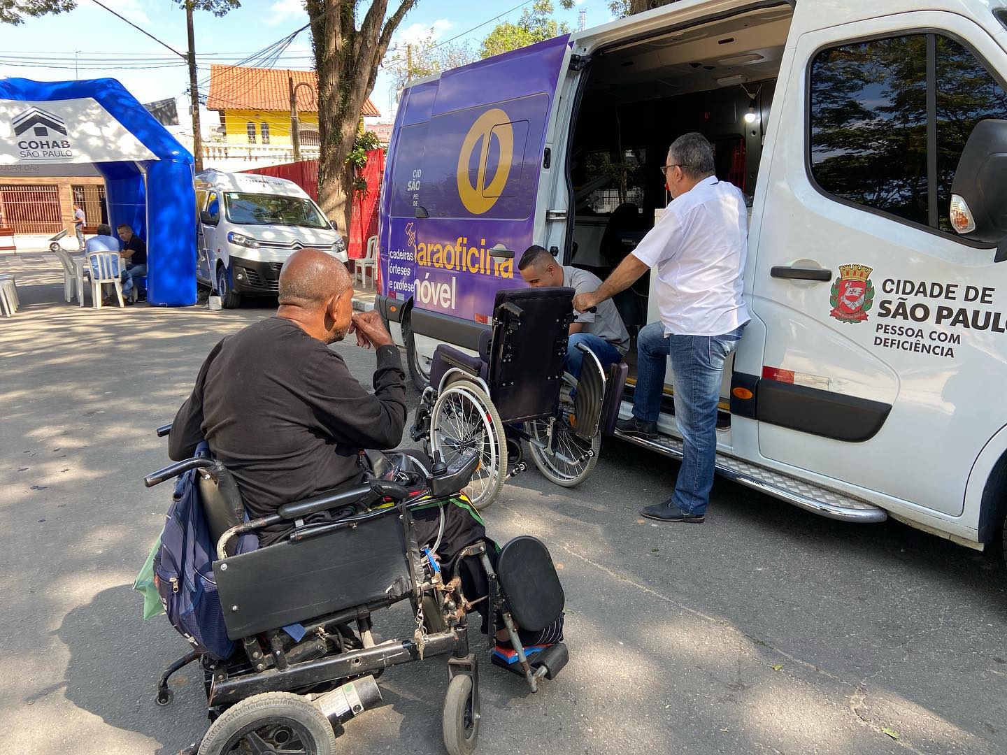 três pessoas na foto: na Paraoficina Móvel técnico realiza manutenção de uma cadeira de rodas, munícipe observa.