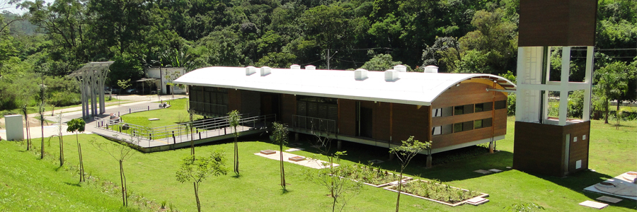 Fotografia da nova sede do Parque Natural Municipal Fazenda do Carmo. Ao centro, está o prédio com paredes de madeira plástica na cor marrom, telhado côncavo na cor branca com claraboias para a entrada da luz natural e rampa na entrada. Em frente ao prédio estão: uma pequena horta dividida em dois quadrantes e dez mudas de árvores jovens. À esquerda estão os paineis fotovoltaicos para captação de energia solar e à direita está um reservatório de água em formato de prédio retangular, também de maderira plástica na cor marrom. Ao fundo, está a vegetação do Parque, composta por árvores de copas verdes. Todo o solo é uma área ajardinada.