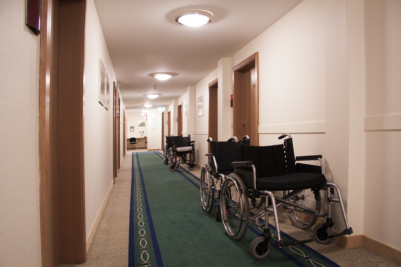 Foto de ambiente. Em um corredor com tapete verde com listras azuis estão quatro cadeiras de rodas apoiadas em uma parede branca. O corredor tem portas marrons dos dois lados. Não tem nenhuma pessoa na imagem.