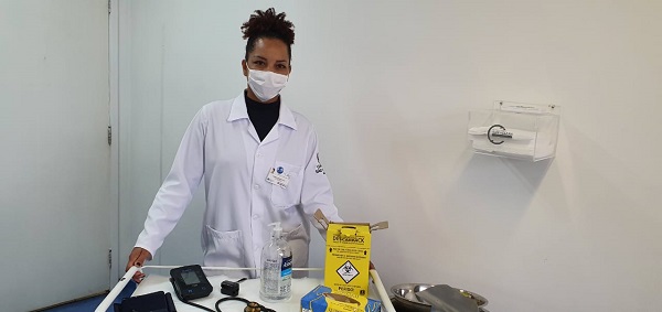 #PraCegoVer: Enfermeira vestindo um jaleco branco, e usando máscara. Sorrindo perto de uma bandeja com álcool em gel, aparelhos de medir pressão e algodão. 