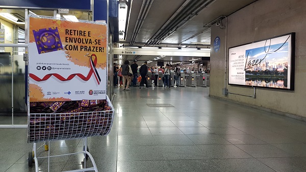 #PraCegoVer: Display de camisinhas está posicionado no canto esquerdo da foto e ao fundo há pessoas próximas às catracas da estação. 