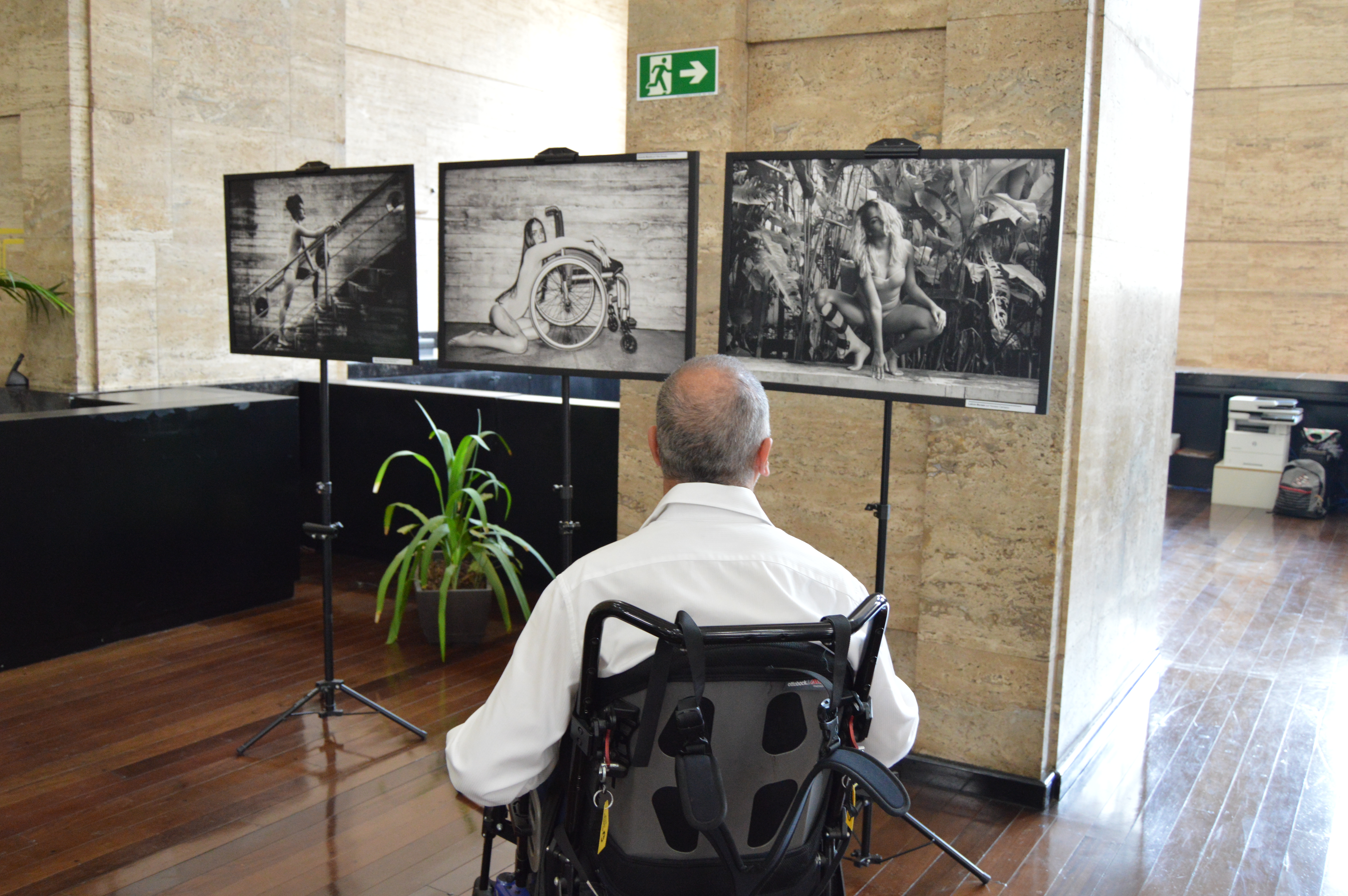 Um homem em uma cadeira de rodas motorizada olhando para fotos em preto e branco de mulheres com deficiência, da exposição Femina Aurea