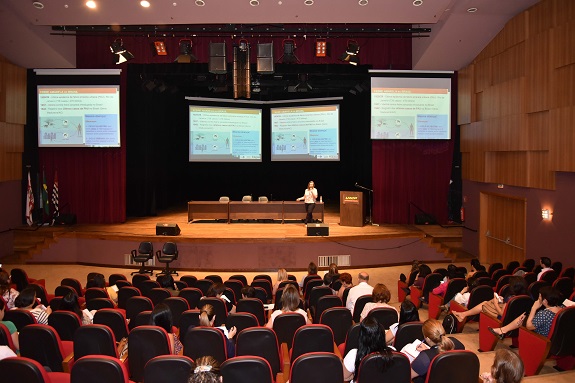 Na foto, pessoas sentadas de costas num auditório, e a palestrante em cima do palco com os slides abertos.