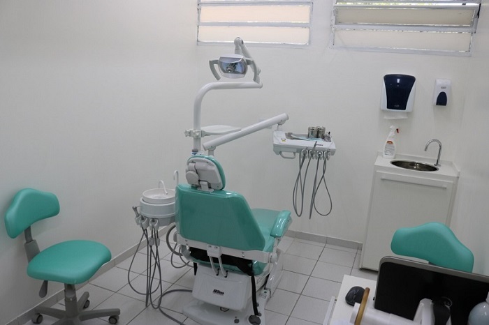 #PraCegoVer: Fotografia da sala de dentista. Ela tem as paredes brancas, e as cadeiras tem o estofado verde. Também há uma pia.
