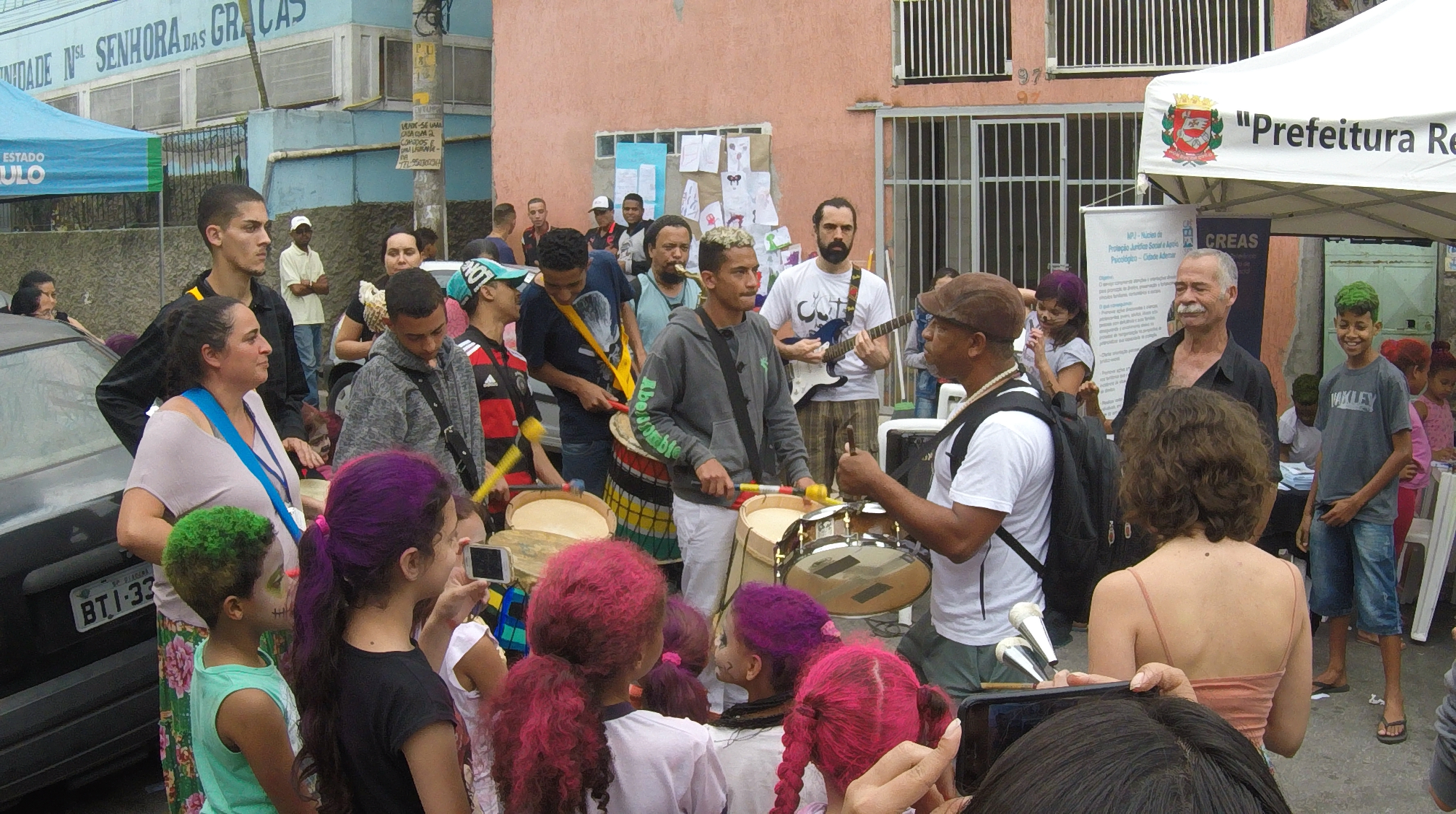 Na rua, no bairro Jardim Miriam, pessoas da comunidade se concentram para ver grupo tocando percussão