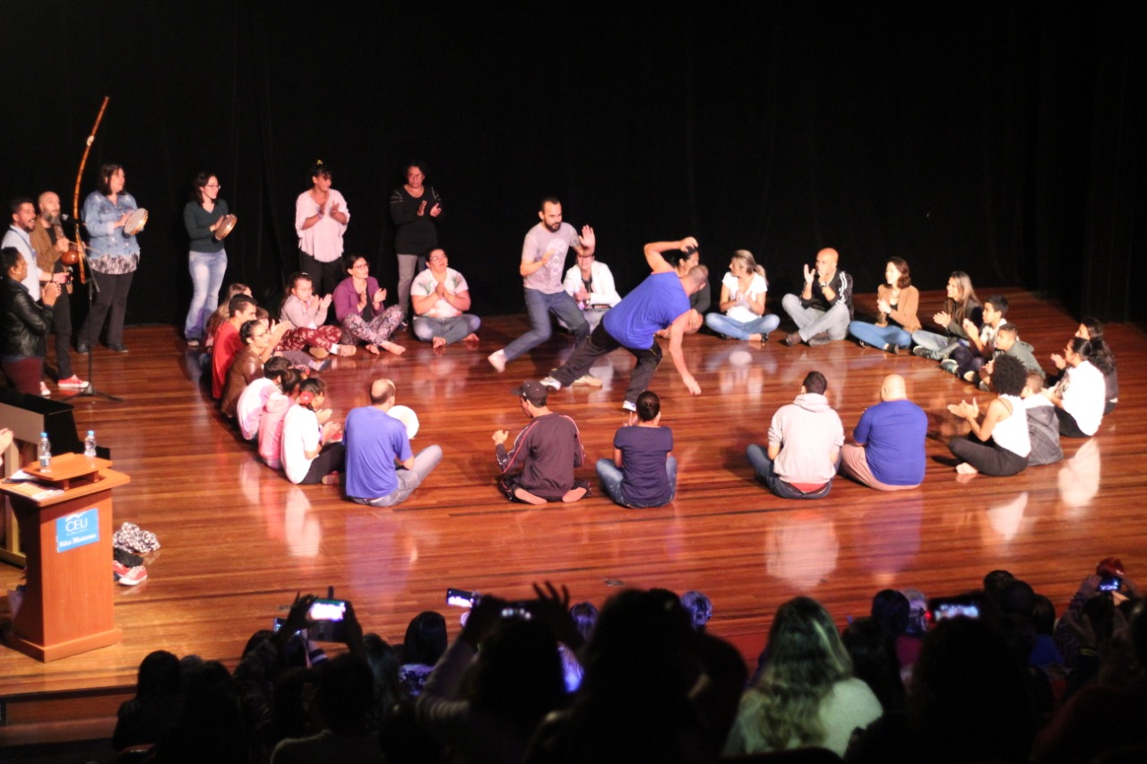 Em cima de um palco, jovens fazem capoeira no meio de uma roda de pessoas