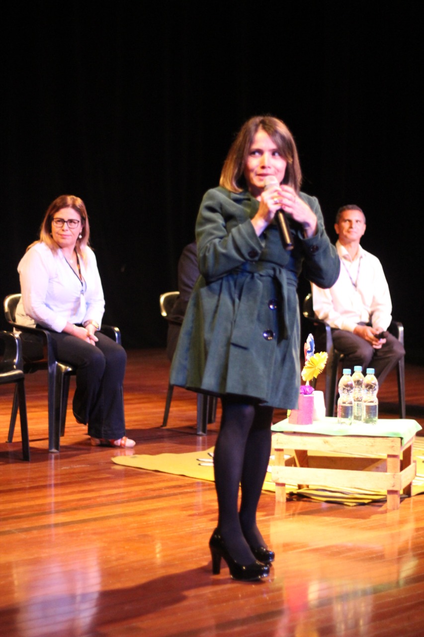 Uma mulher em pé discursa com o microfone enquanto outras pessoas estão sentadas no palco