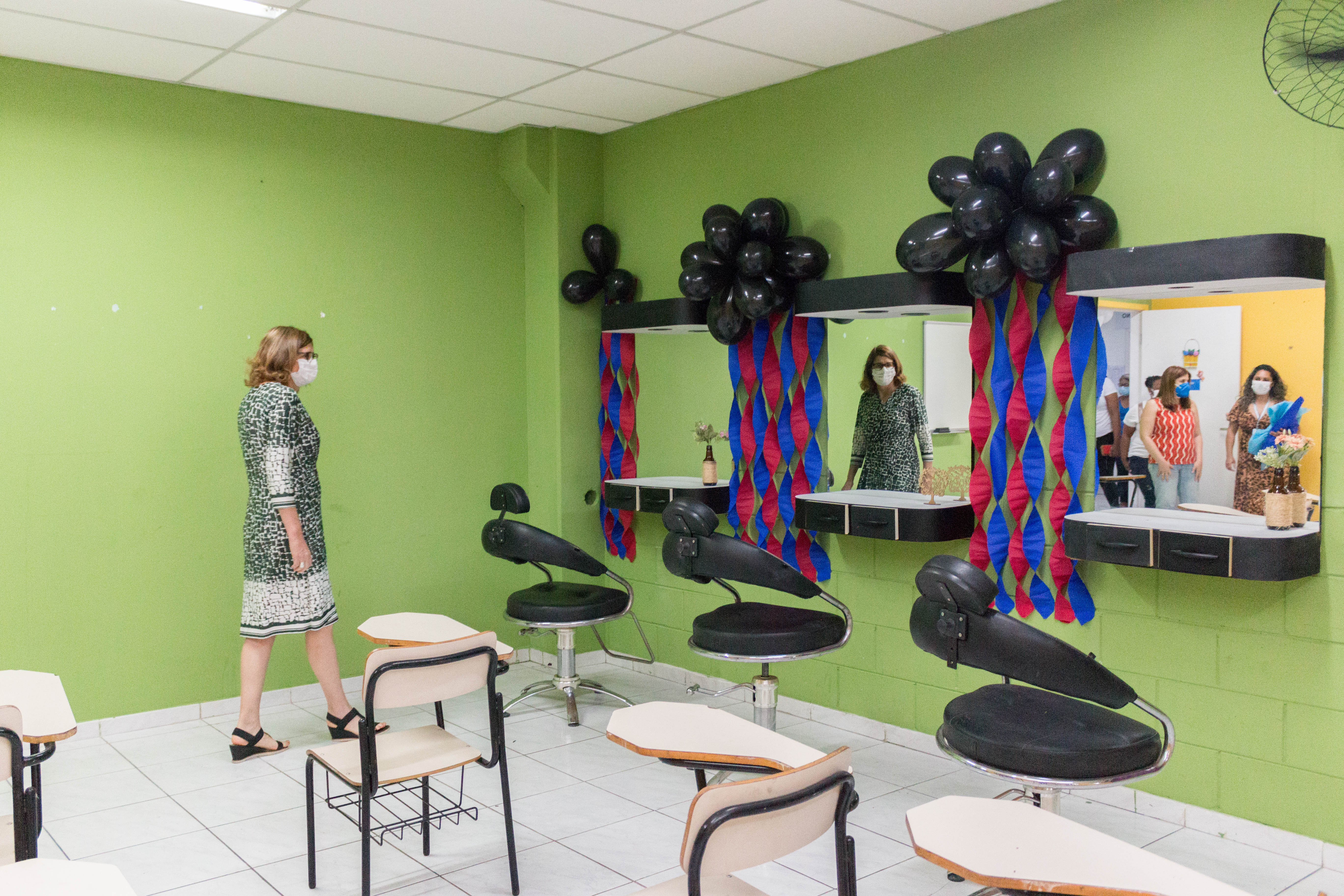 A secretária Berenice Giannella adentrando uma sala com paredes verdes, há algumas mesas escolares e na lateral três espelhos e cadeiras de cabeleireiro.