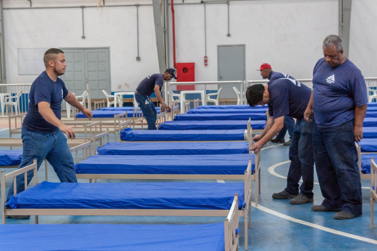Cinco homens de uniforme azul marinho trabalham em sala branca na organização de camas brancas com colchões azuis.