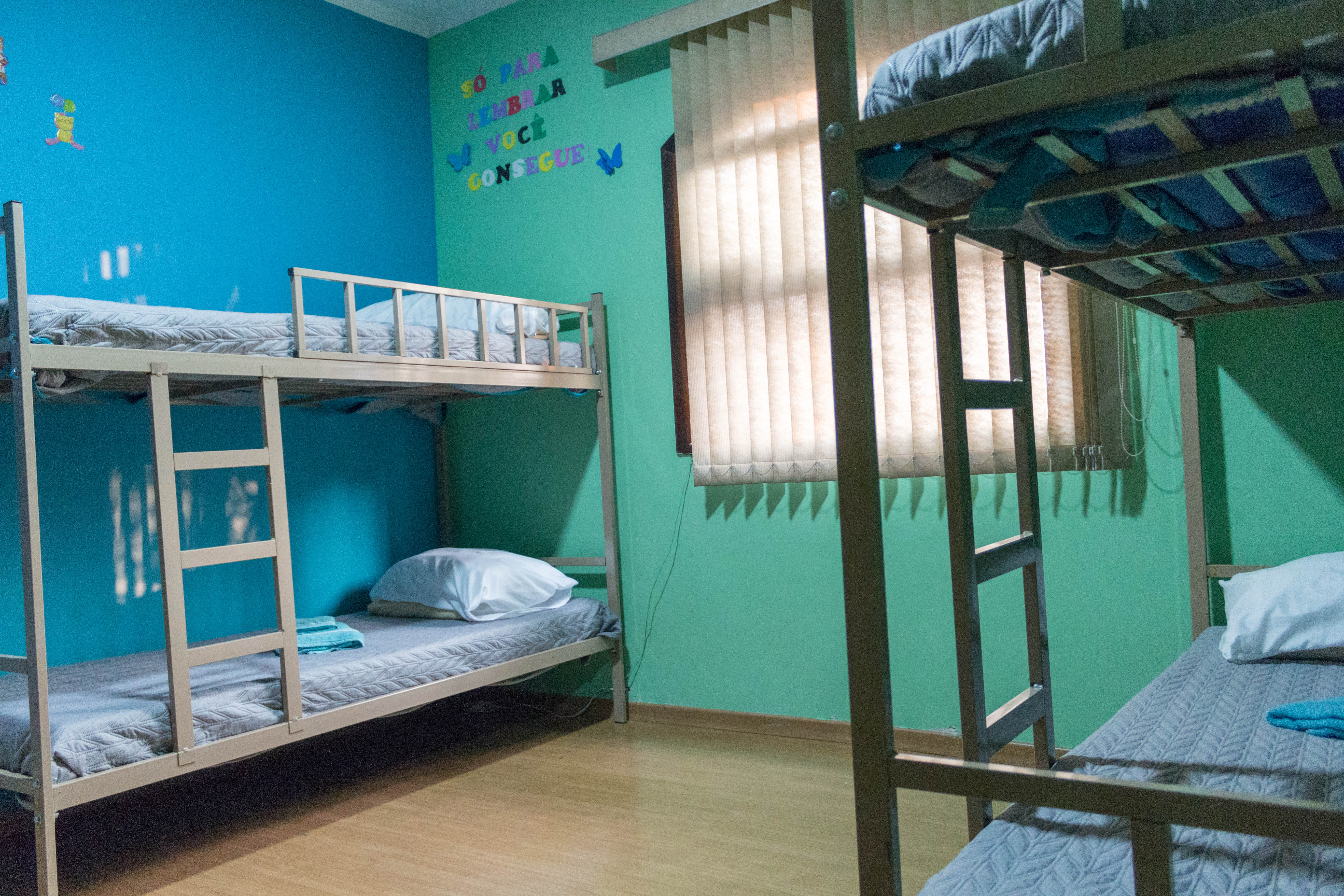 Quarto com uma parede verde e outra azul contém dois beliches. No canto, há a mensagem “Só para lembrar você consegue” no canto e ao centro há uma janela fechada por cortinas brancas.