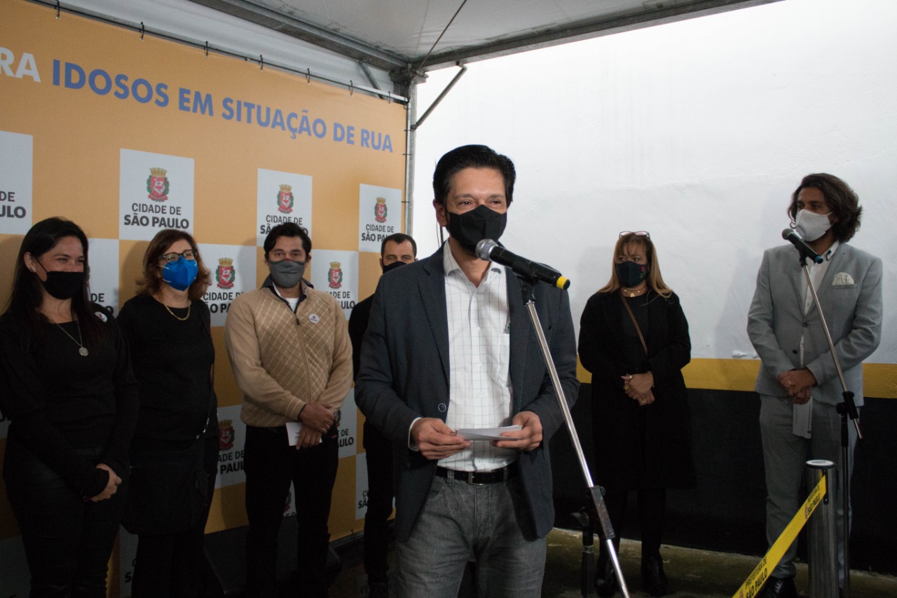 Prefeito Ricardo Nunes em cerimônia oficial de inauguração em frente a um painel quadriculado em amarelo e branco, com brasões da prefeitura de São Paulo intercalados de forma repetitiva.