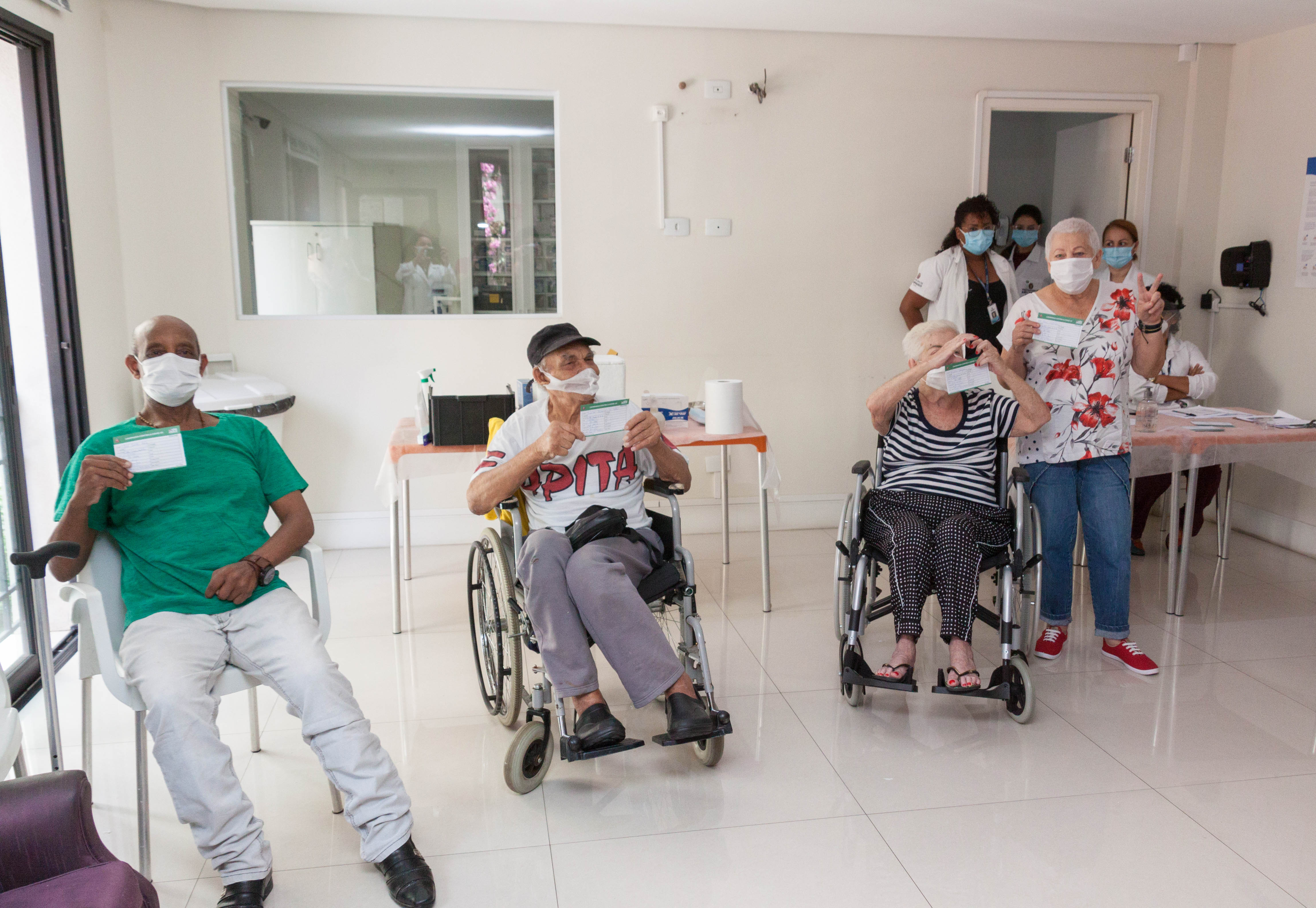 Quatro idosos sendo eles dois homens e duas mulheres posam para foto erguendo seus respectivos cartões de vacinação. Dos quatro, apenas uma senhora está em pé e os demais sentados. Todos usam máscaras brancas descartáveis e estão a frente de uma mesa que contém a vacina CoronaVac.