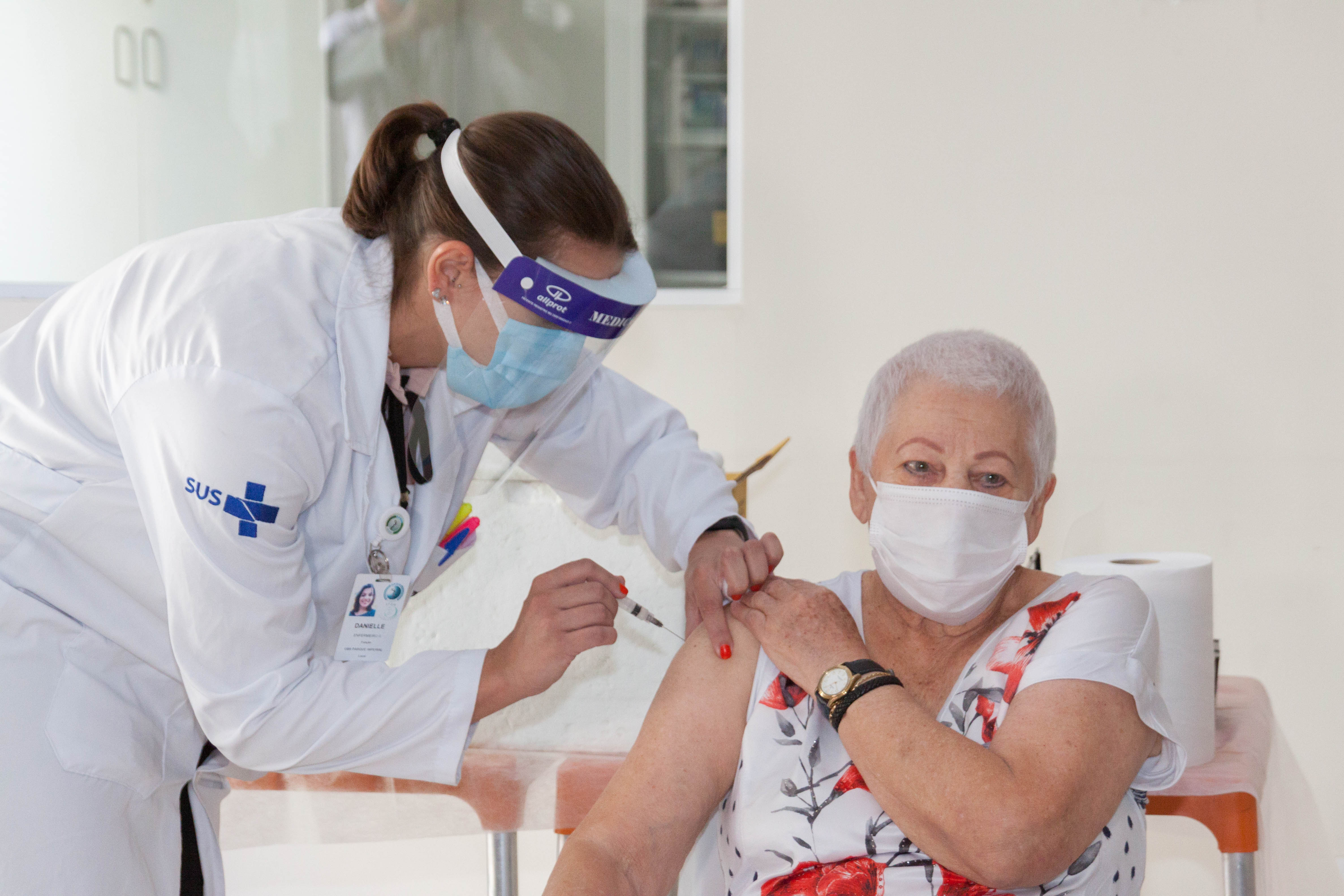 A convivente da Instituição de Longa Permanência para Idosos Vila Mariana, Rosa Nullman, em seu momento de vacinação. A vacina é aplicada por uma enfermeira vestindo os devidos aparatos de segurança.