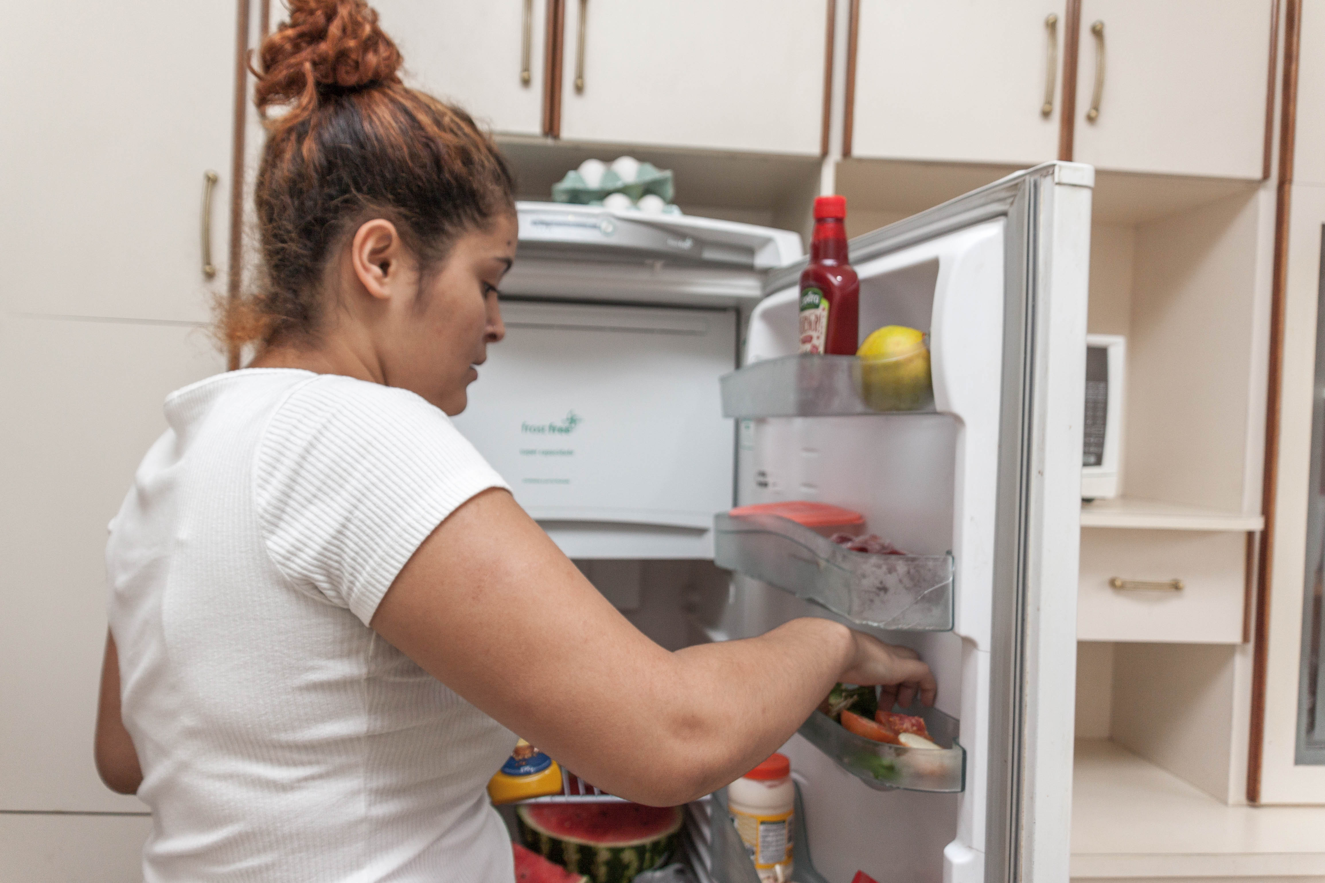 Moradora de República Jovem está arrumando alimentos na porta da geladeira. Ela está usando uma camiseta branca e os cabelos, em tons ruivos, estão presos em um coque.