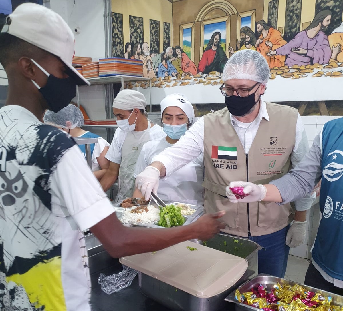 O secretário Carlos Bezerra Junior durante a ação “Islam contra a Fome” servindo comida na bandeja que é segurada por um homem acolhido do Complexo Boracea.