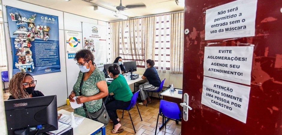 Ambiente interno da sala de atendimento do Centro de Referência de Assistência Social (CRAS). A sala possui três mesas de atendimento, em primeiro plano, uma senhora está sendo atendida em pé e mais duas outras pessoas são atendidas em outras duas mesas. Uma janela ao fundo está aberta. O ambiente tem ventilador de teto de pás, chão de tacos de madeira e cadeiras tipo concha plástica azul. A porta de entrada da sala é vermelha e tem três avisos impressos em papel A4 na horizontal com orientações sobre uso de máscara, evitar aglomerações e entrada de uma pessoa por vez.