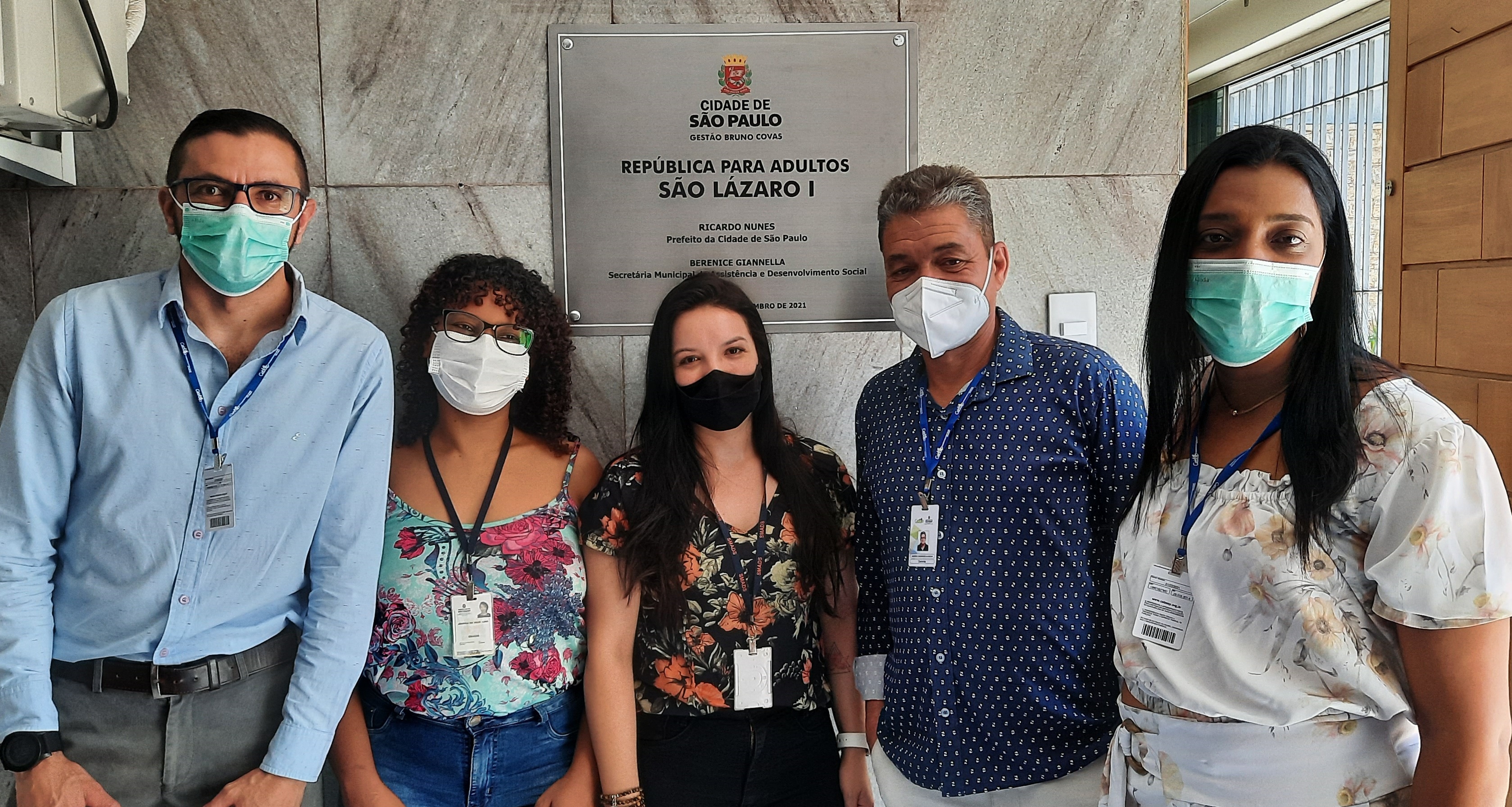 Em frente à placa de inauguração da República para Adultos São Lázaro I, funcionários da rede direta de assistência social posam para foto.