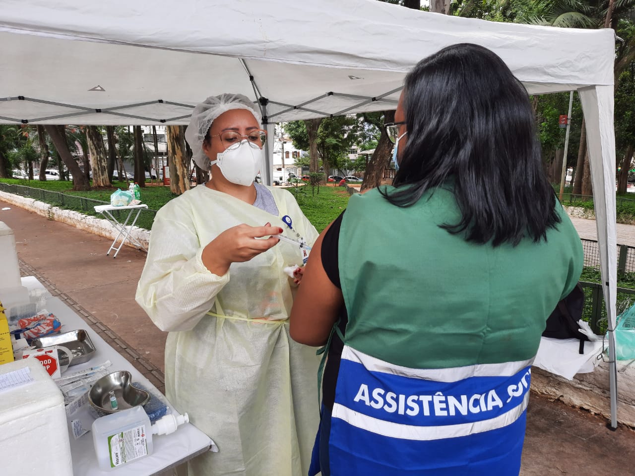 Sob tenda branca e ao lado de uma mesa com diversos utensílios médicos, enfermeira vestindo todos os equipamentos de proteção individual aplica a vacina em uma orientada que veste o colete da Assistência Social.